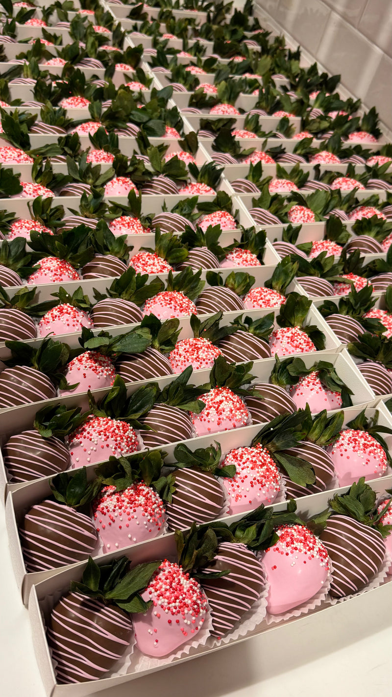 Rows of assorted chocolate-dipped strawberries arranged in white boxes, including milk chocolate strawberries with pink drizzle and pink chocolate strawberries topped with red, pink and white sprinkles.